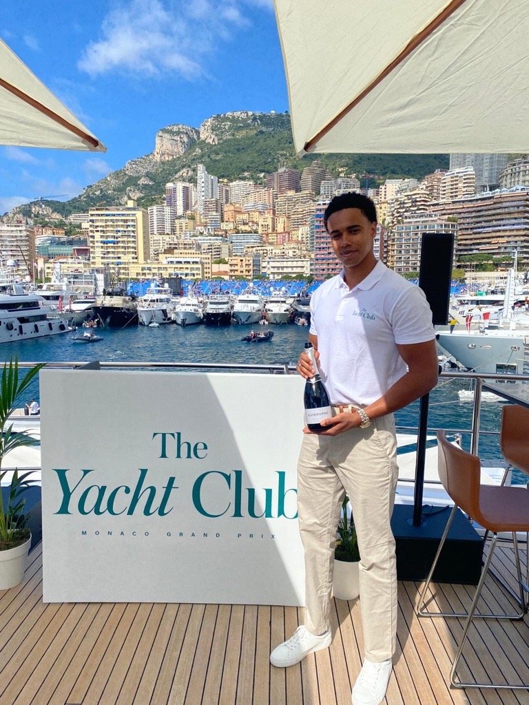 Young man in white polo and beige pants holding a bottle at Monaco Yacht Club, with boats and hillside buildings in the background.
