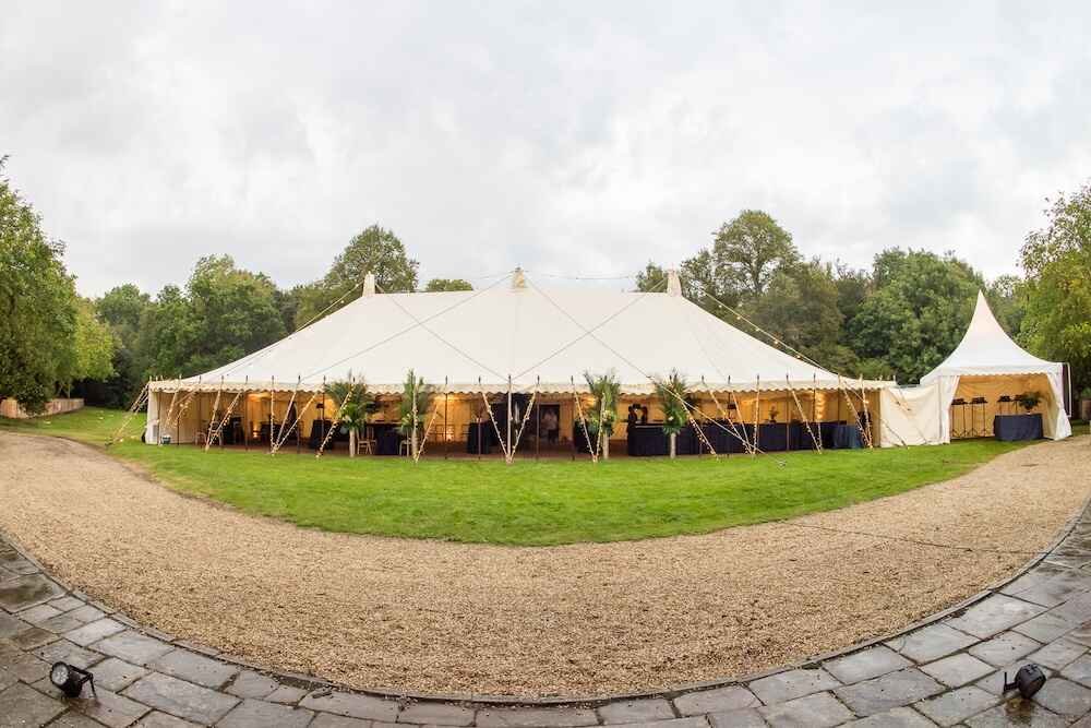 Large outdoor event tent set up on grassy area with trees in background, surrounded by gravel paths.