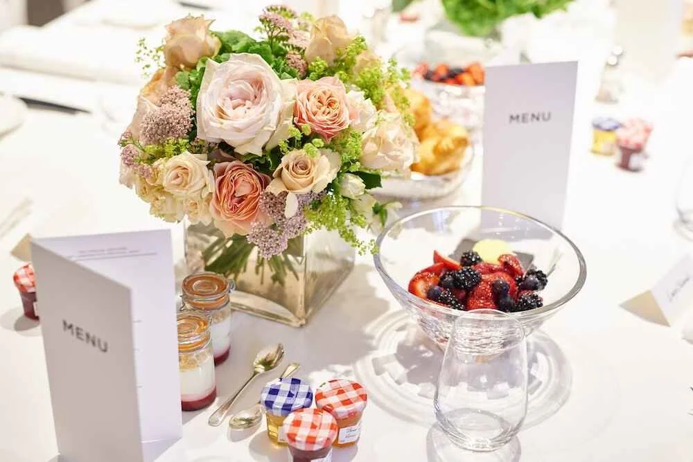 A table with a floral centerpiece of pink and cream roses, a bowl of mixed berries, small jars of yogurt and jam, and a menu card.