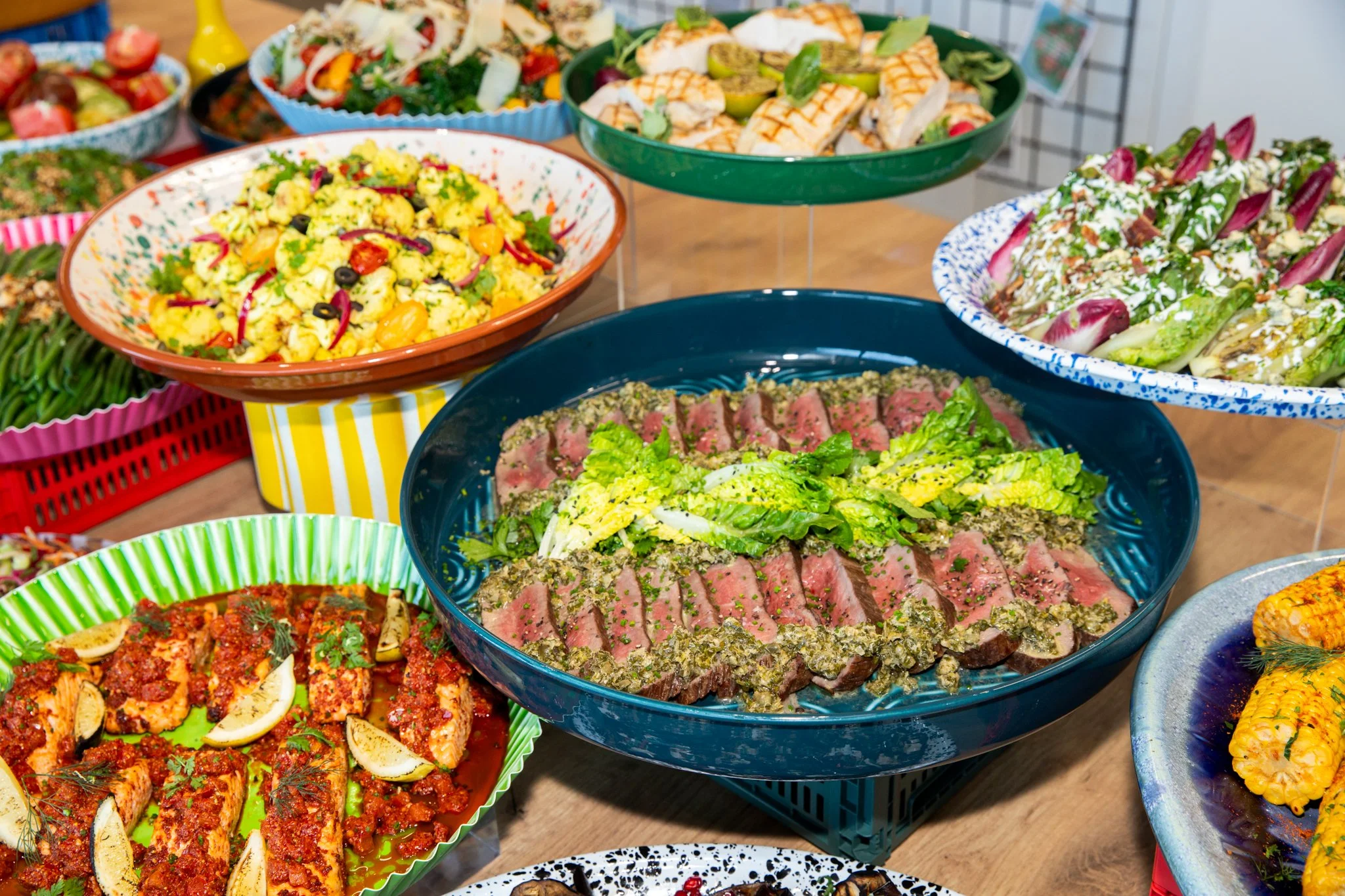 Assorted colorful salads and grilled meats displayed on a table, including beef, chicken, and vegetable dishes in various bowls and plates.