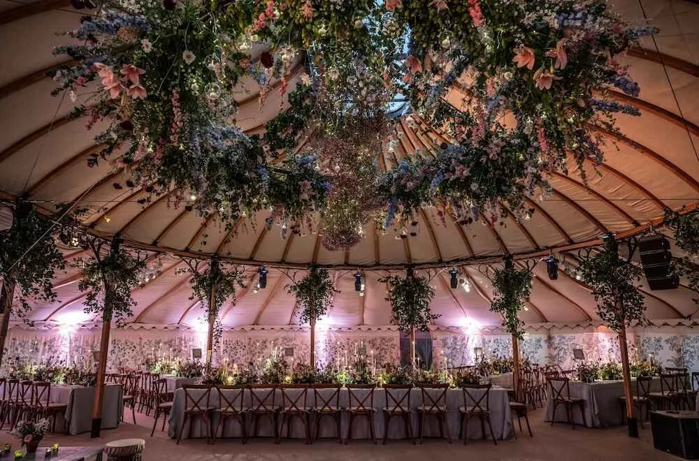 Wedding reception tent decorated with hanging flowers and greenery, with long tables covered in white tablecloths and surrounded by wooden chairs, pink and purple lighting creating a romantic atmosphere.