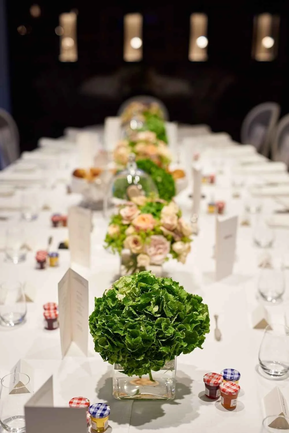 Elegant banquet table decorated with a green leafy centerpiece, surrounded by pink and white floral arrangements, place cards, glasses, and utensils, set for a formal event.