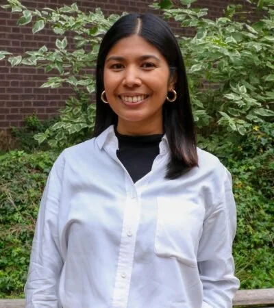 A young woman with shoulder-length dark hair, wearing a white button-up shirt and gold hoop earrings, standing outdoors in front of green foliage and a brick wall, smiling at the camera.