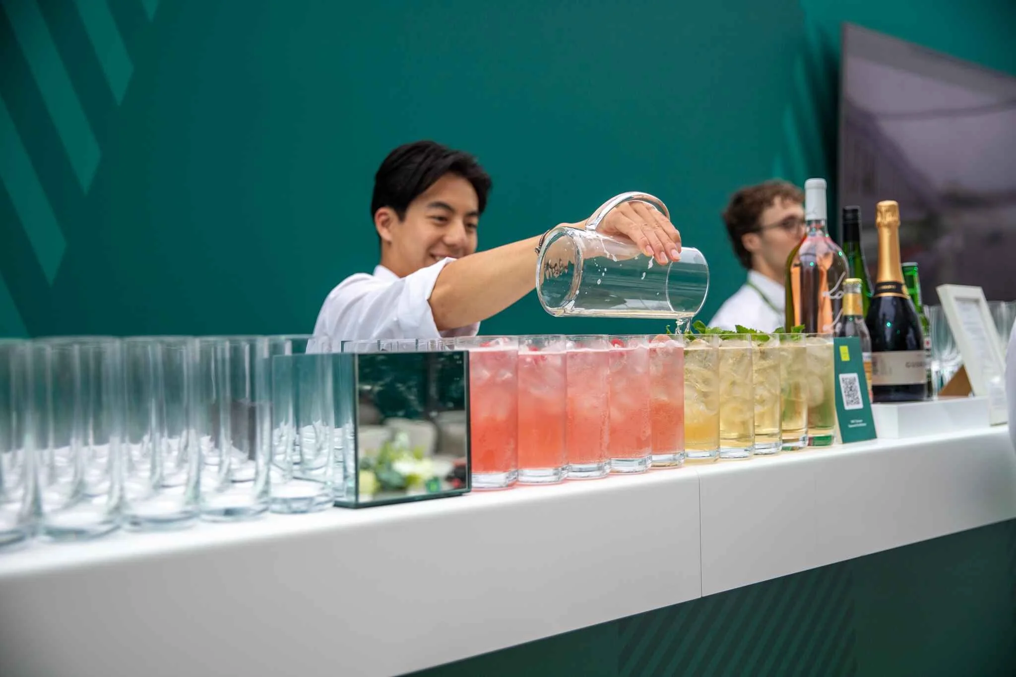 A bartender pouring a drink from a glass pitcher into a colorful glass at a bar, with bottles of champagne and other alcohol on the counter.