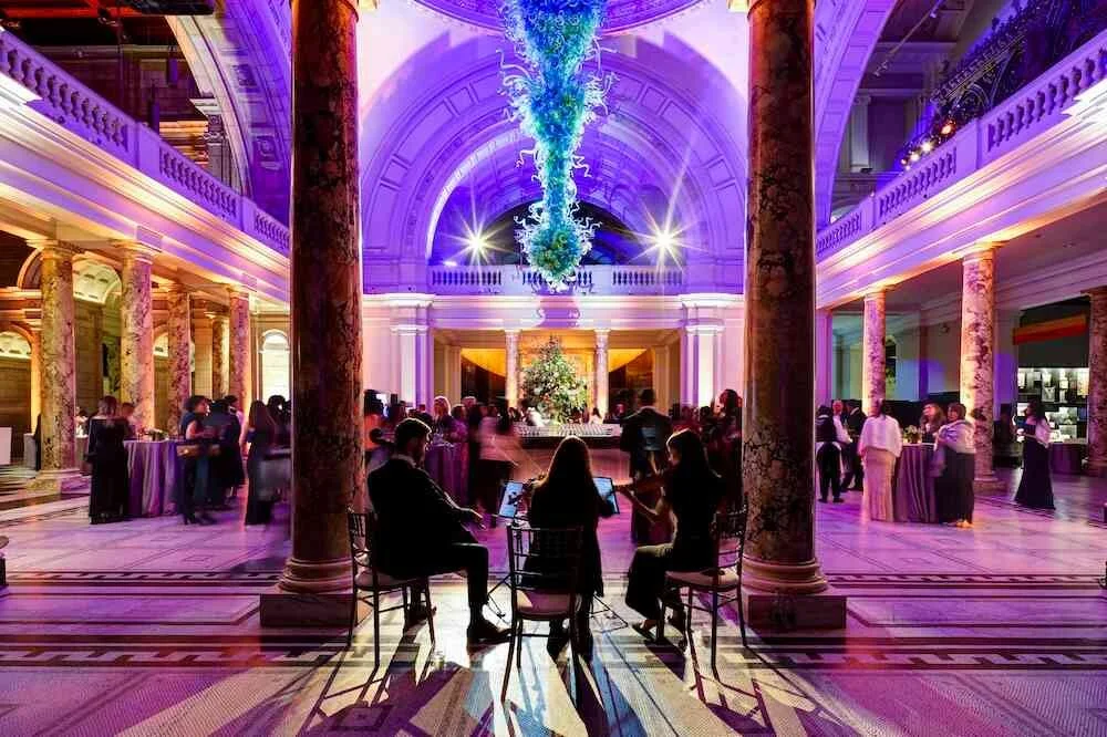 Elegant indoor event in a grand hall with purple and pink lighting, large columns, a chandelier, and people socializing and dining at tables.