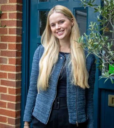 A young woman with long blonde hair smiling outdoors, standing in front of a brick wall and blue door, with a green plant nearby.