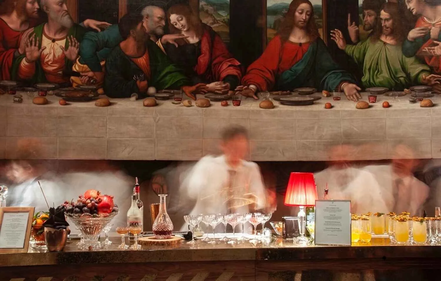 A large painting of Leonardo da Vinci's 'The Last Supper' on the wall depicts Jesus and his apostles sitting at a long table. Below the painting, a bar with drinks, glasses, and a red lamp is visible, with people blurred in motion in front of the bar.