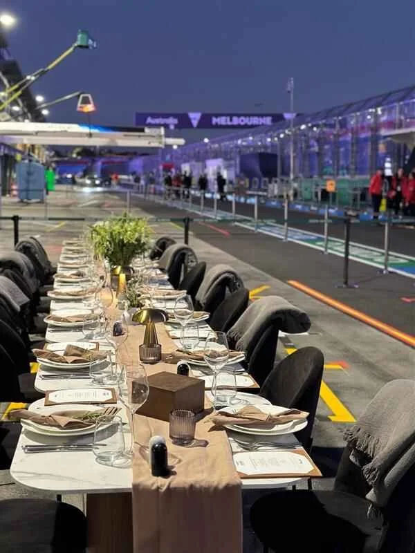 A long dining table set with glasses, plates, napkins, and centerpieces in a racing garage or pit lane at night with a sign reading 'Melbourne' visible in the background.