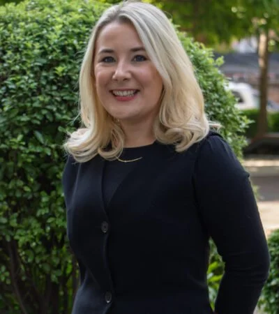 A woman with blonde hair smiling, standing outdoors beside green bushes, wearing a dark blazer.