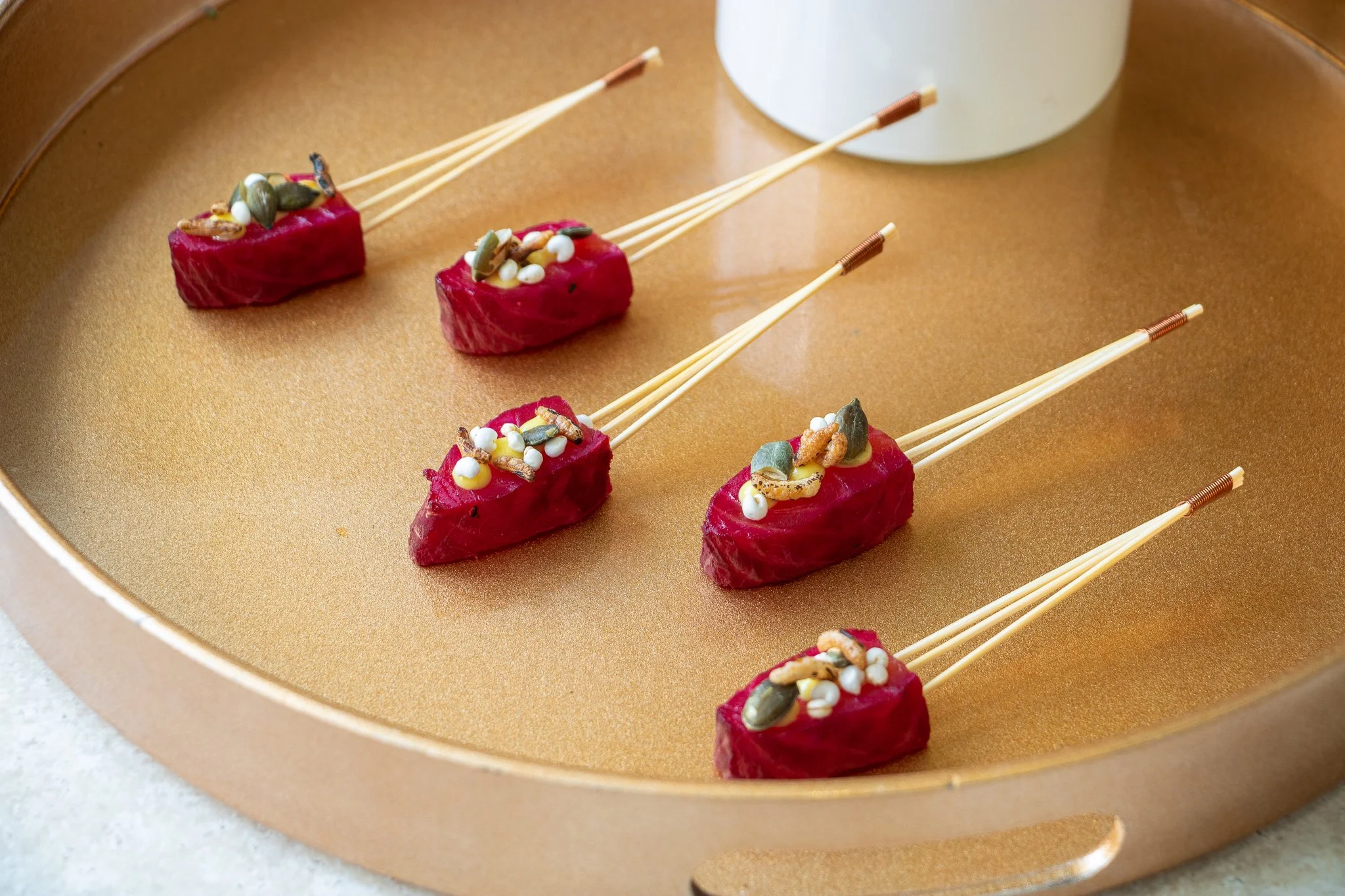 Five pieces of sushi with red fish on a gold tray, garnished with seeds and small toppings, with white cup in the background.