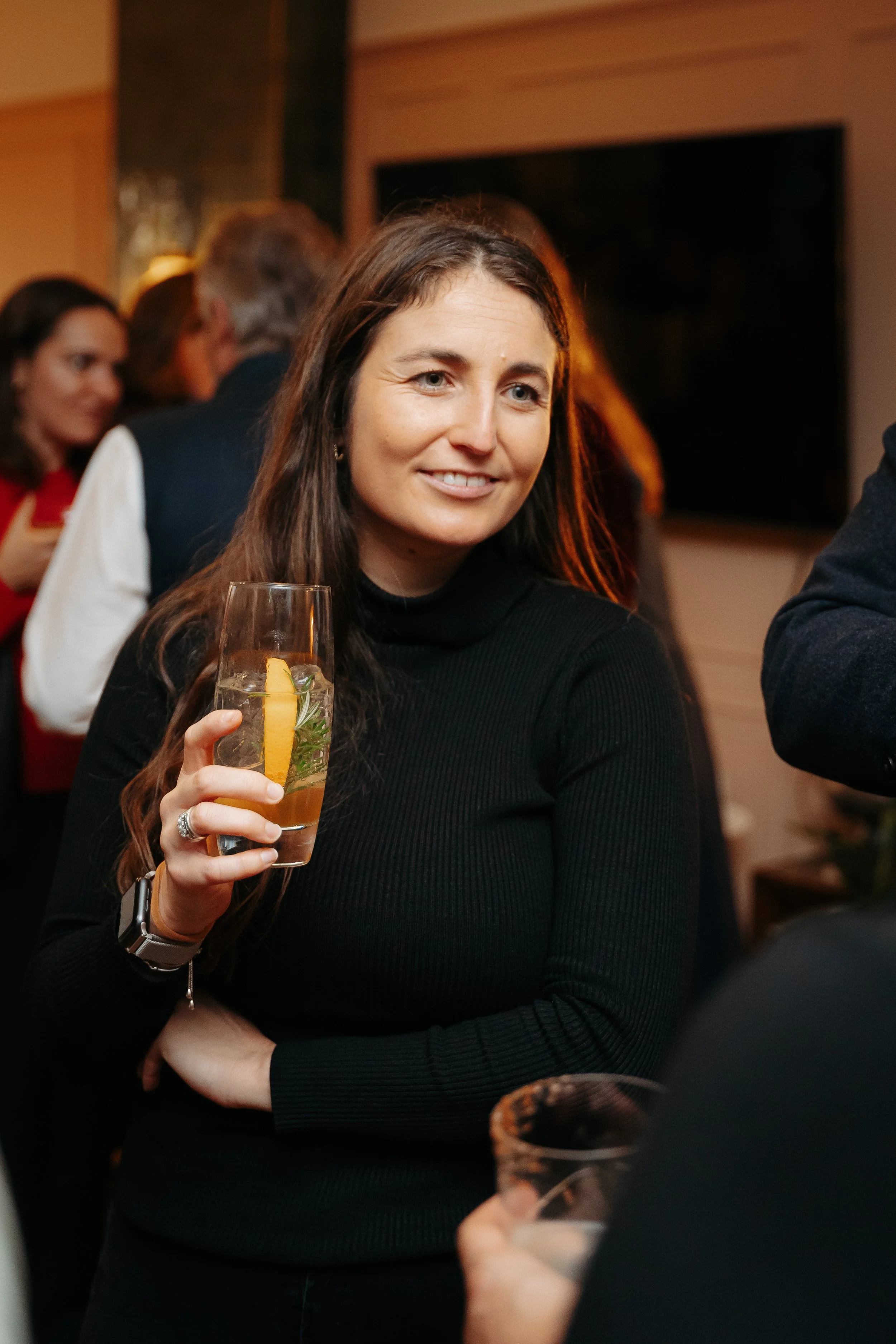 A woman with long, wavy brown hair smiling outside in front of green leafy plants, wearing a light-colored blazer and dark top.
