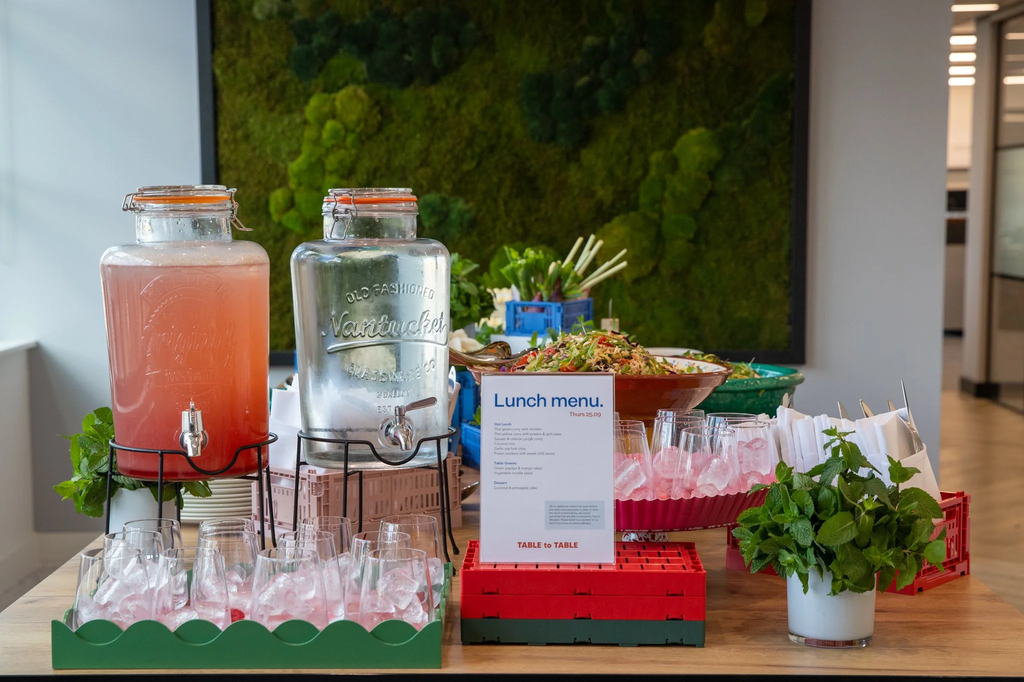 Buffet table with pink and clear drinks in dispensers, salads, and a lunch menu sign in a modern restaurant.