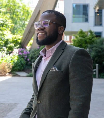 A man in a gray suit with pink shirt and glasses standing outdoors near modern buildings and greenery.