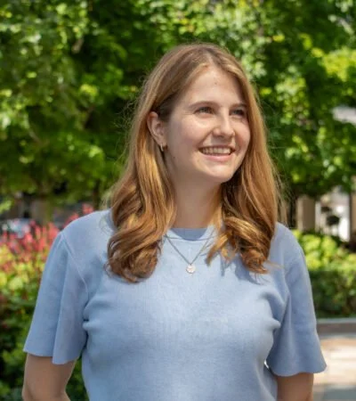 A young woman with light brown hair and a blue shirt smiling outdoors in a park with green trees background.