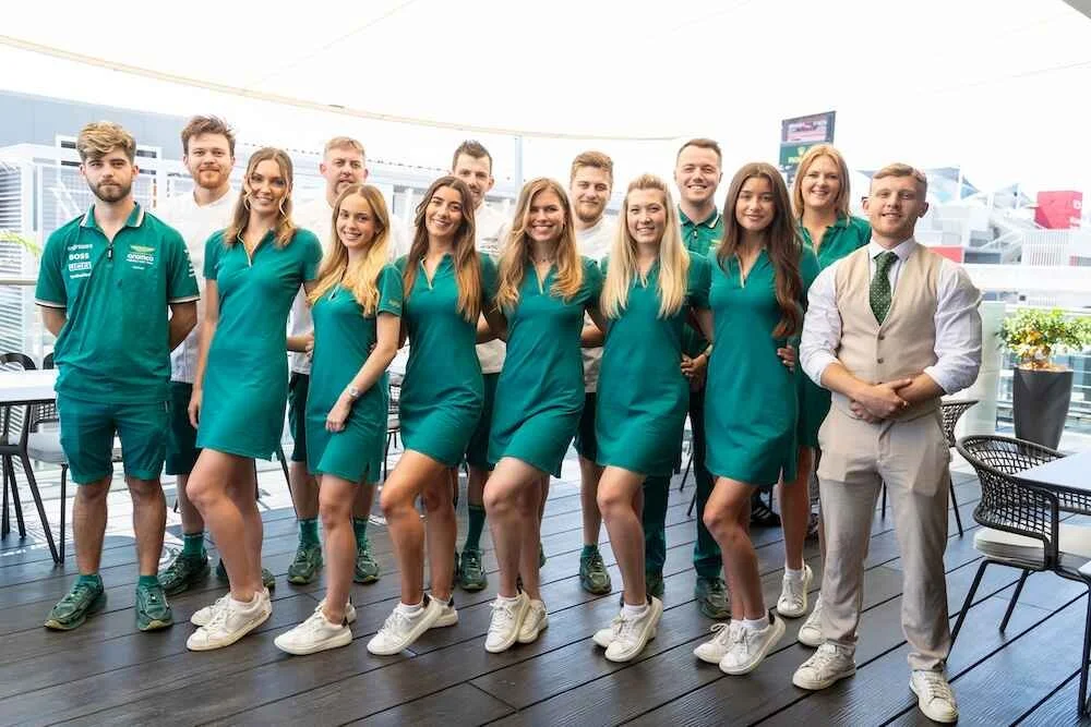 Group of waitstaff members in teal uniforms and a manager in beige vest in a bright indoor space.