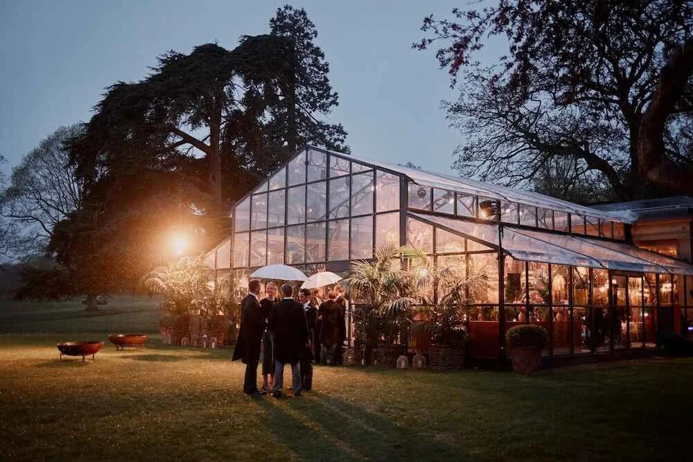 A group of people dressed in formal attire stand outside a large glass greenhouse or conservatory in the evening, illuminated from within, with trees in the background.