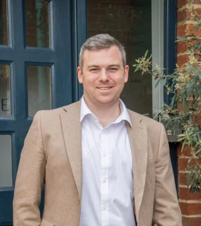 A man with short gray hair wearing a beige blazer and white dress shirt standing outside near a brick building with a blue window frame and some green foliage.