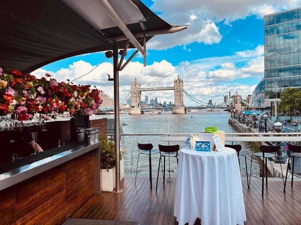 View of Tower Bridge over the River Thames with a restaurant patio in the foreground, featuring a table with a white tablecloth and chairs, and a modern glass building on the right.