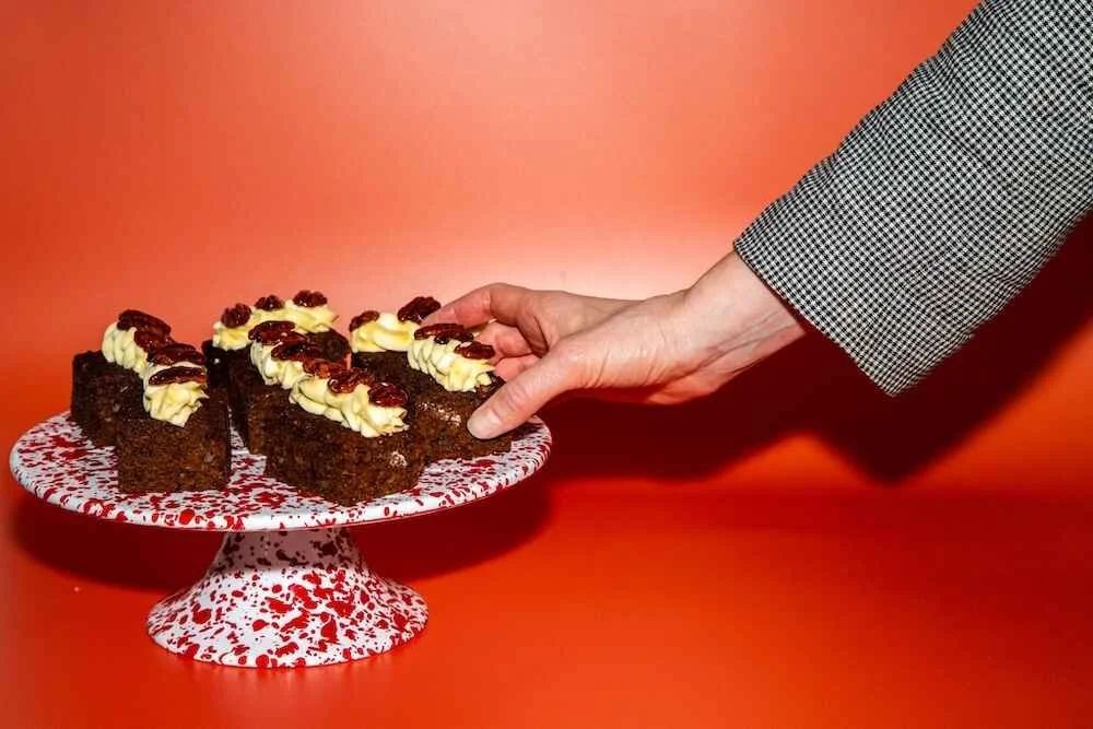 A hand reaching for pieces of chocolate cake with white frosting and red topping on a white cake stand with red splatter pattern, set against an orange background.