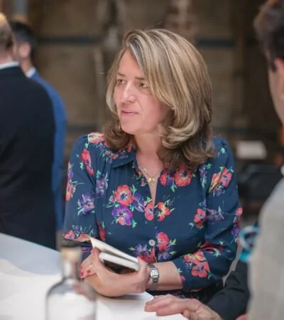 A woman with shoulder-length blonde hair wearing a floral blouse, holding a smartphone, engaging in conversation in a casual social setting.