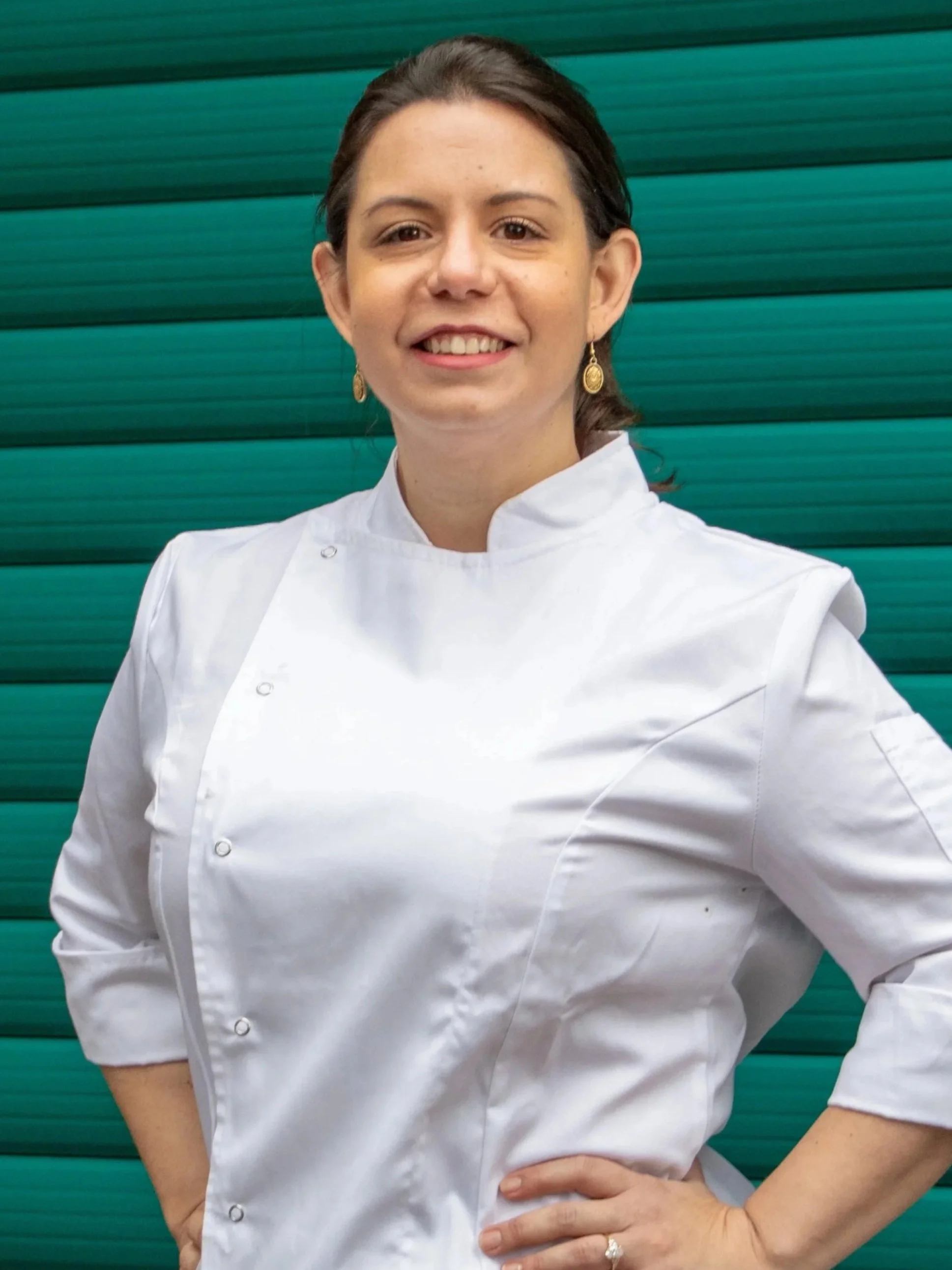 A woman wearing a white chef's coat, standing in front of a green rolling shutter door, smiling with her hands on her hips.
