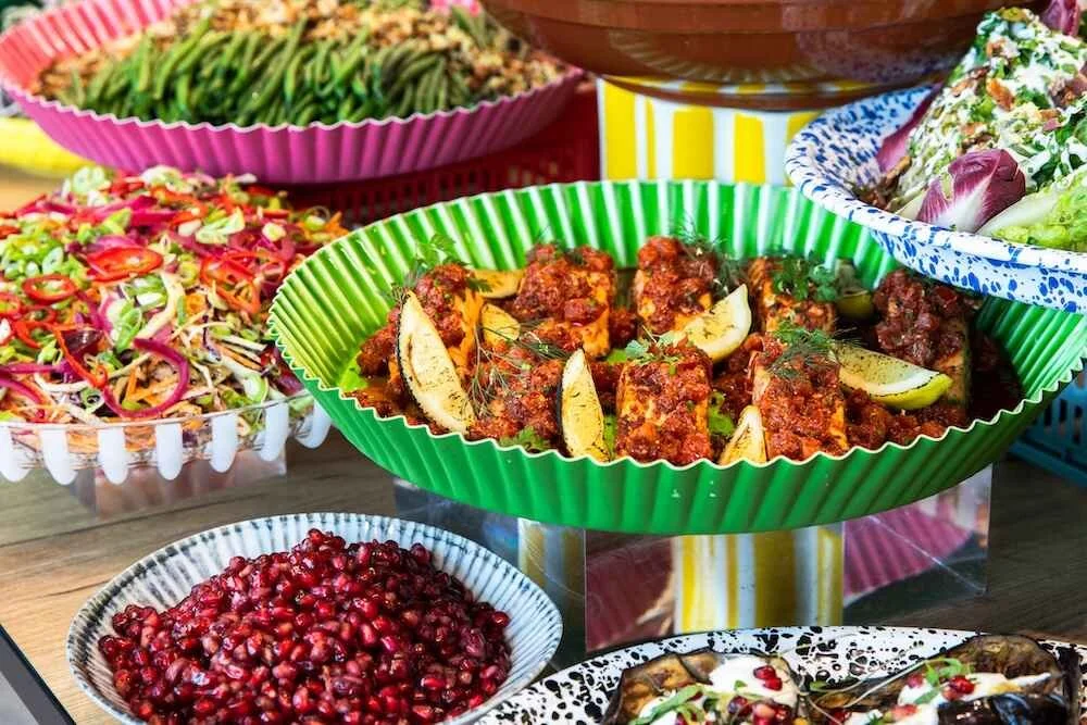 A colorful spread of Mediterranean dishes including a platter of grilled meat with lemon wedges, a bowl of pomegranate seeds, a salad with sliced vegetables, and other assorted salads on decorative plates and bowls.