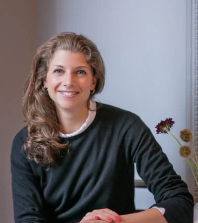 A woman with long, curly brown hair, smiling, sitting indoors near a window with a flower arrangement nearby.