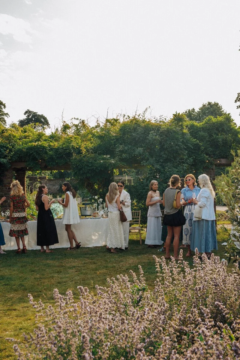 Group of women socializing outdoors at a garden party or wedding reception, standing near a table with food and drinks, surrounded by greenery and flowering plants.