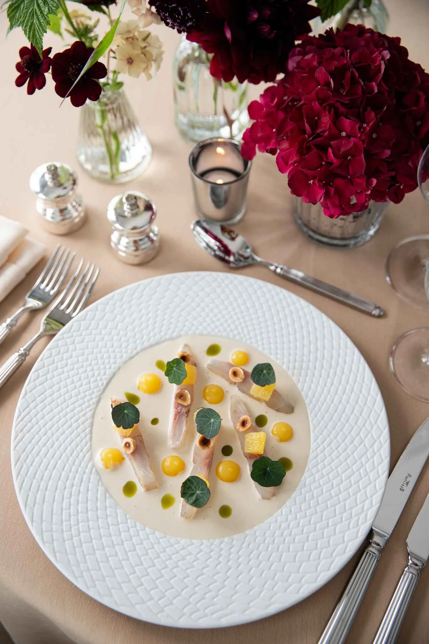 Elegant table setting with a white textured plate holding a sashimi dish garnished with yellow spheres, green leaves, and small nuts, surrounded by silverware, candles, and red and white floral arrangements.