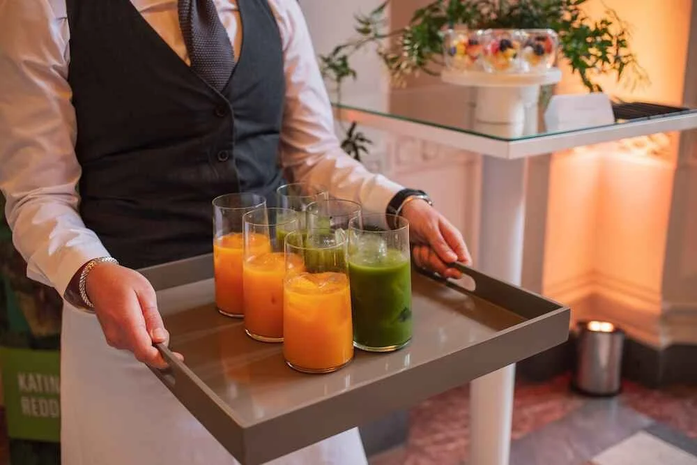 Person in formal attire carrying a tray with six colorful drinks, possibly smoothies or juices, in a well-lit indoor setting with plants and decorative lighting in the background.