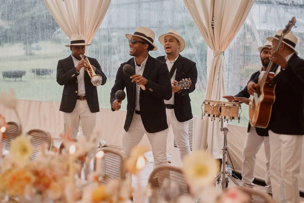 A live band performing at an event inside a tent. The band members are dressed in black blazers, white pants, and hats, with one holding maracas, another with a guitar, and others playing percussion instruments. The tent has curtains and large windows, with a blurred floral arrangement in the foreground.