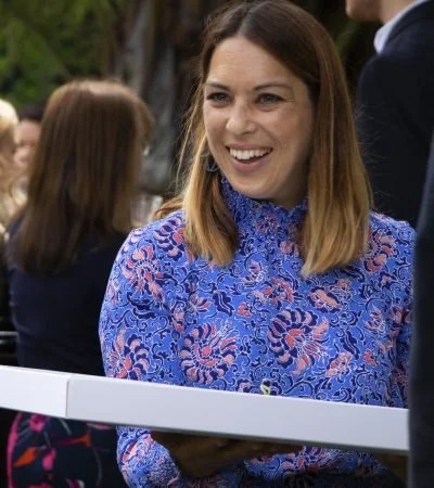 A woman with shoulder-length brown hair smiling and holding a white tray at an outdoor event.