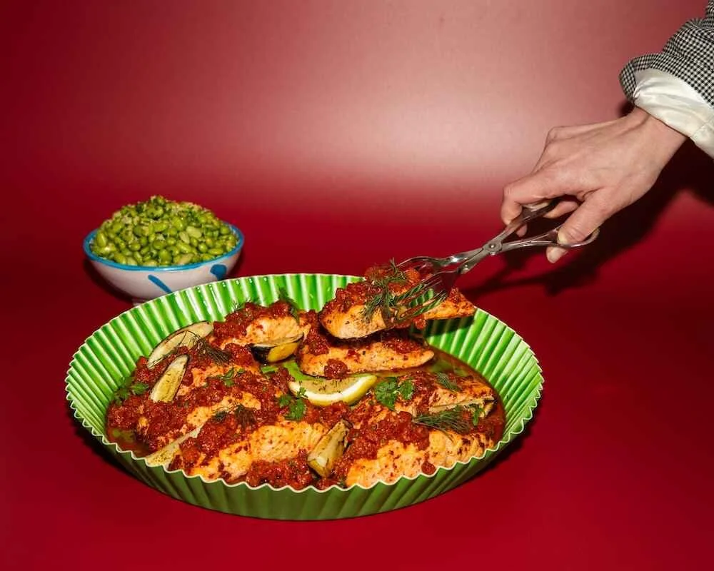 Hand serving baked chicken with tomato sauce from a green dish, with a bowl of green peas in the background.