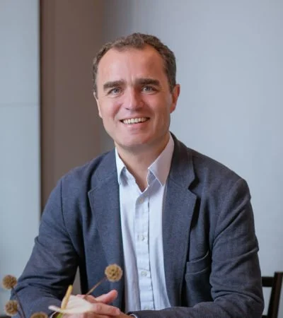 A smiling man in a gray blazer and white shirt sitting at a table in an indoor setting.