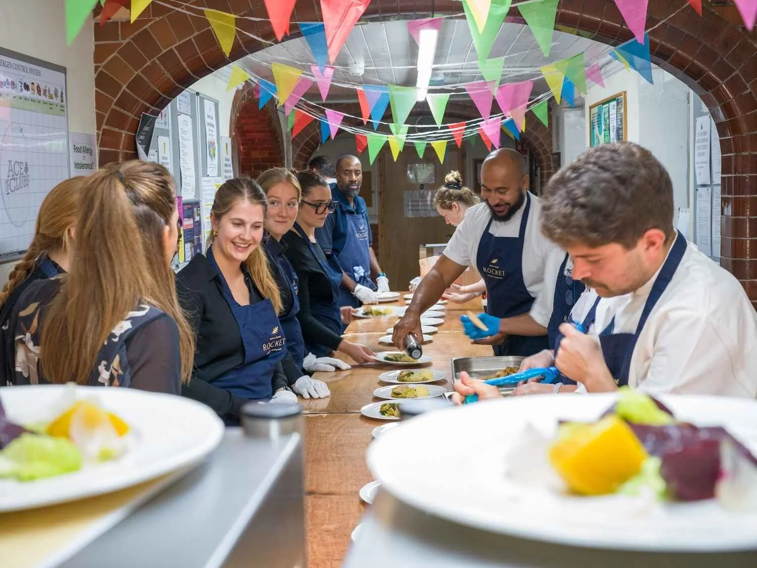 A group of people, some wearing blue aprons with a logo, participating in a cooking class or food event under colorful pennant banners in a brick-walled room, showcasing plated dishes on a long counter.
