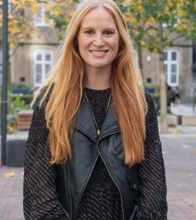 A young woman with long red hair smiling outdoors, wearing a black leather vest over a black sweater, with trees and buildings in the background.