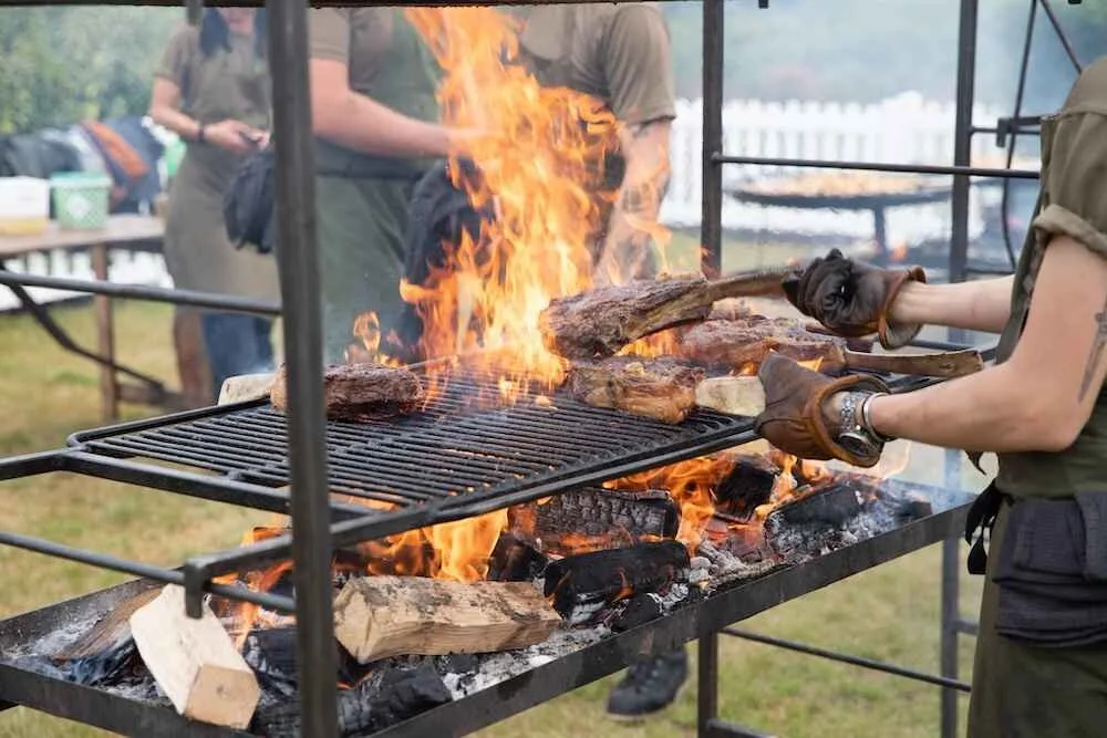 Person wearing gloves and a watch grilling large pieces of meat over an open flame at an outdoor event.