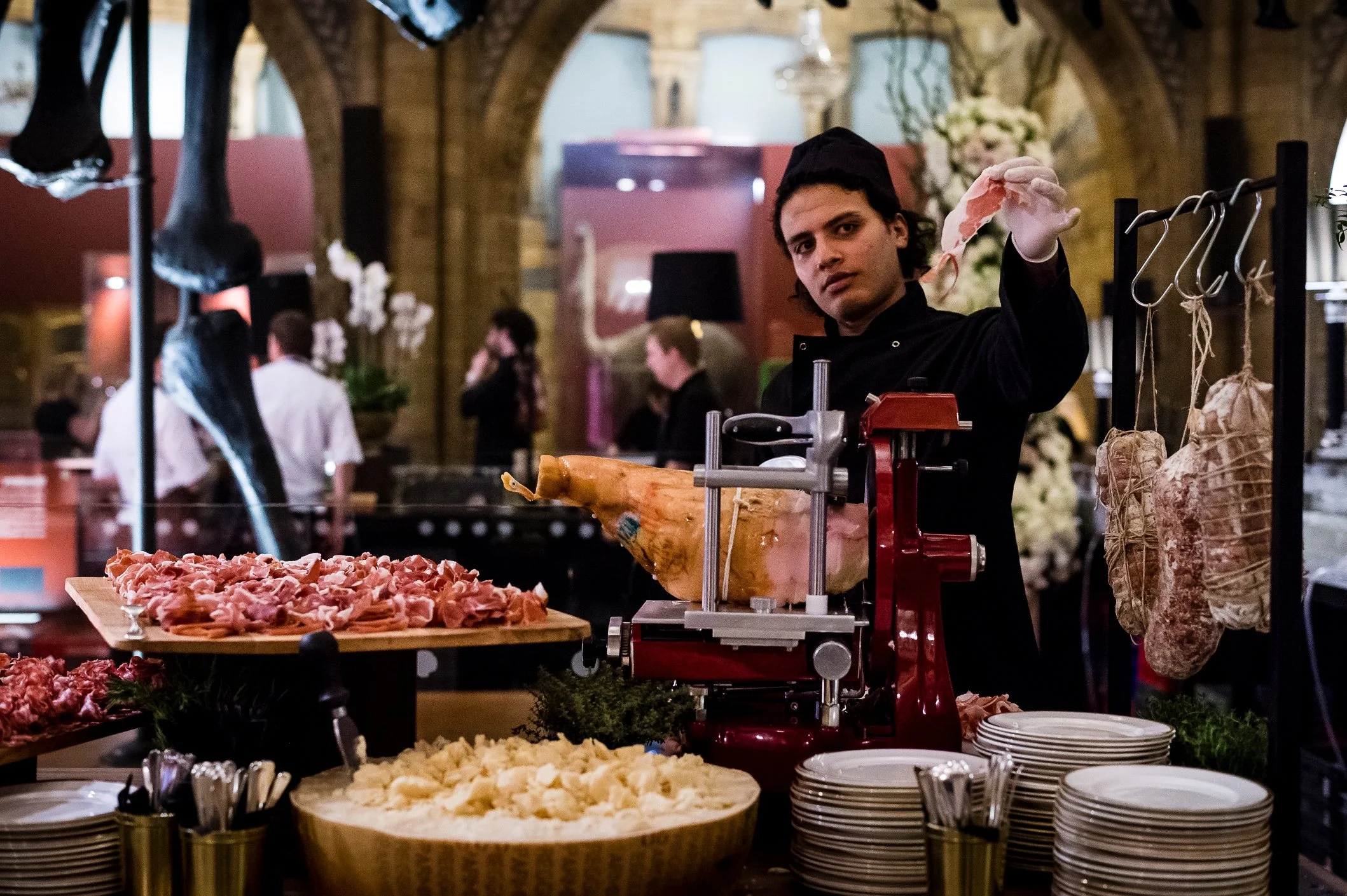 A man at a butcher shop or deli slicing meat from a large leg of ham with a meat slicer. The counter displays various cuts of meat, and plates are stacked nearby. Hanging on the right side are sausages and charcuterie. In the background, there are shoppers and flowers in a warmly lit setting.