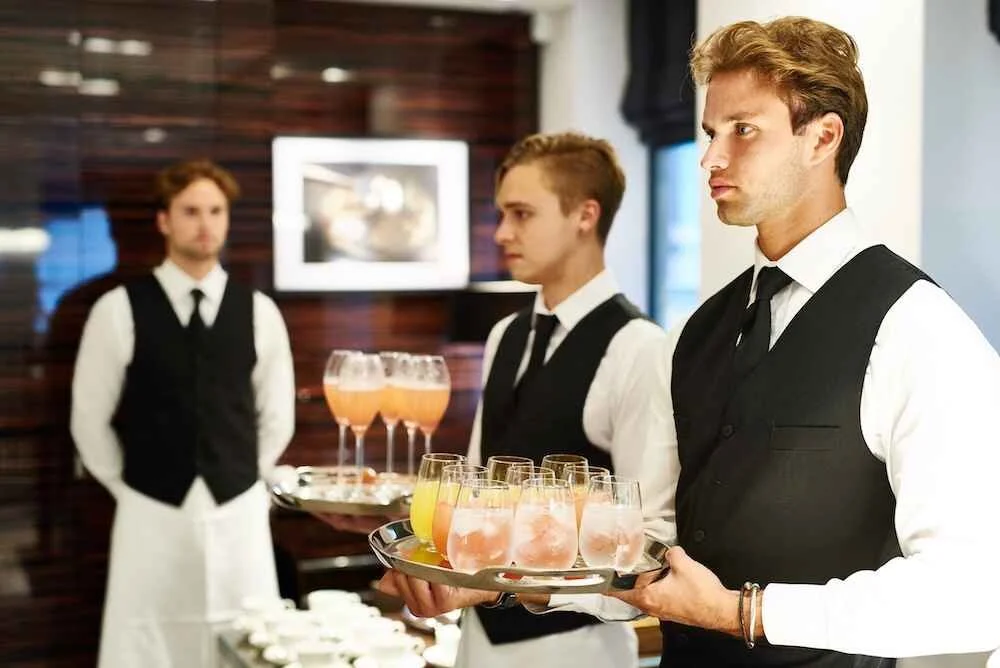 Three young male waiters in white shirts, black vests, and black ties standing in a restaurant, holding trays with drinks, with a wooden wall and a framed picture in the background.