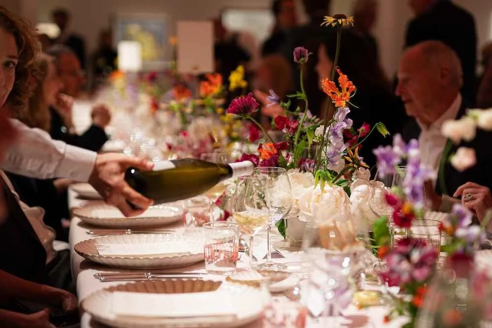 People at a long banquet table with floral centerpieces, being poured wine, in a warmly lit setting.
