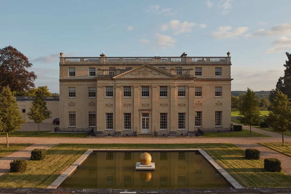 A large, historic, beige mansion with columns and decorative cornices, surrounded by trees, with a reflective pond in the foreground and a blue sky with scattered clouds above.