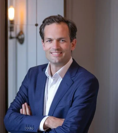 A man in a blue suit standing with arms crossed in an indoor setting, smiling at the camera.