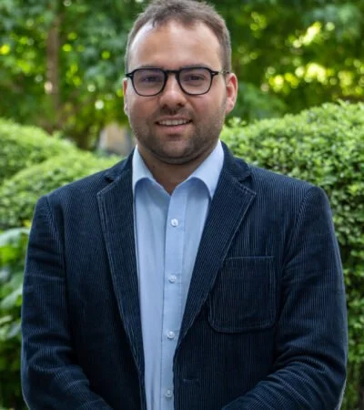 A man with glasses in a navy blazer and light blue shirt standing outdoors with greenery in the background.