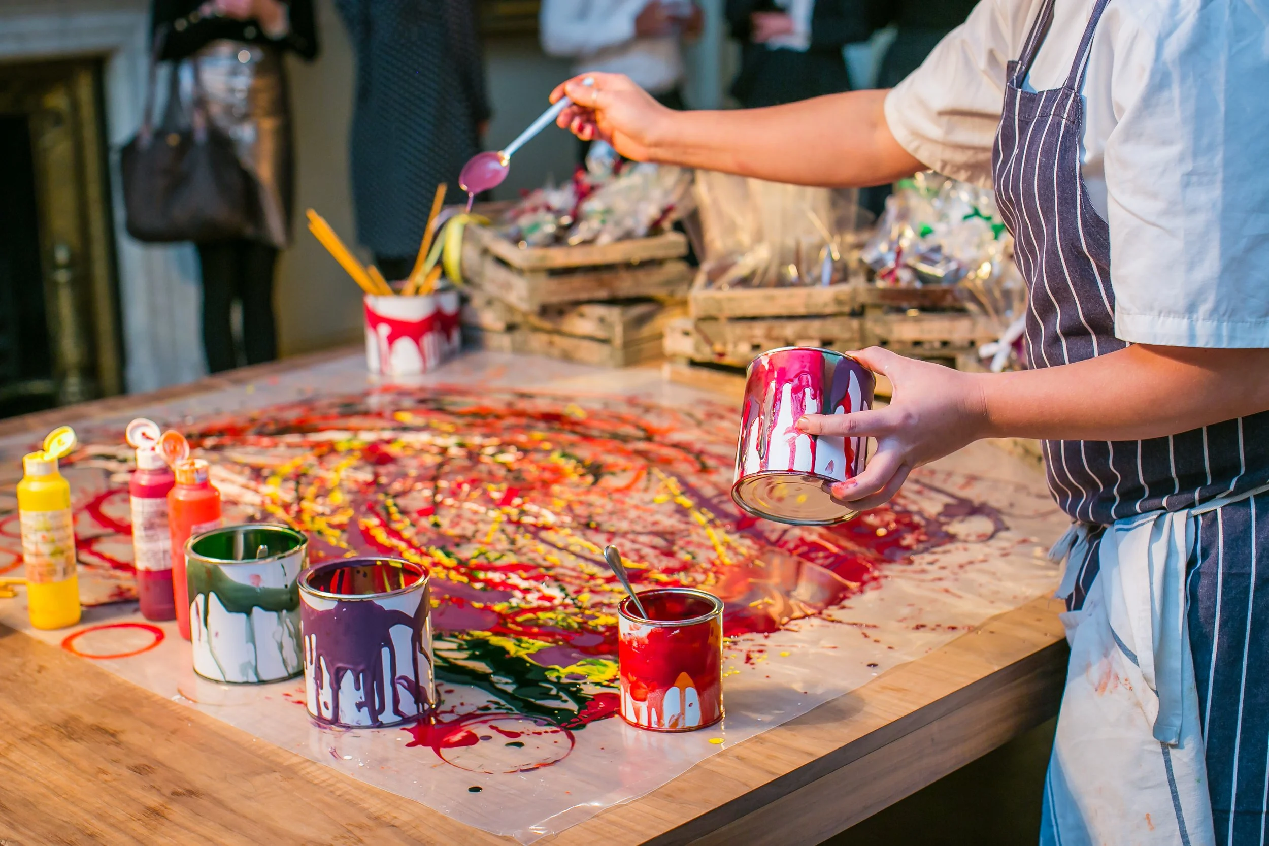 Person creating a colorful abstract painting on a table with various paint cans, brushes, and paints.