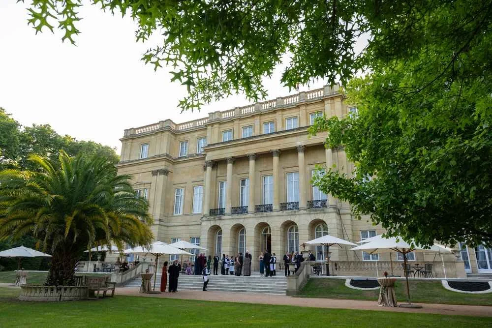 A large, elegant historic building with tall columns and multiple stories, situated in a lush green garden with palm trees, bushes, and umbrellas providing shade. People are gathered on the steps and around the entrance, suggests a formal event or gathering.