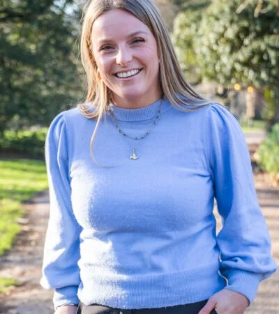 A young woman with long blonde hair smiling outdoors, wearing a blue sweater and a necklace, with trees and sunlight in the background.