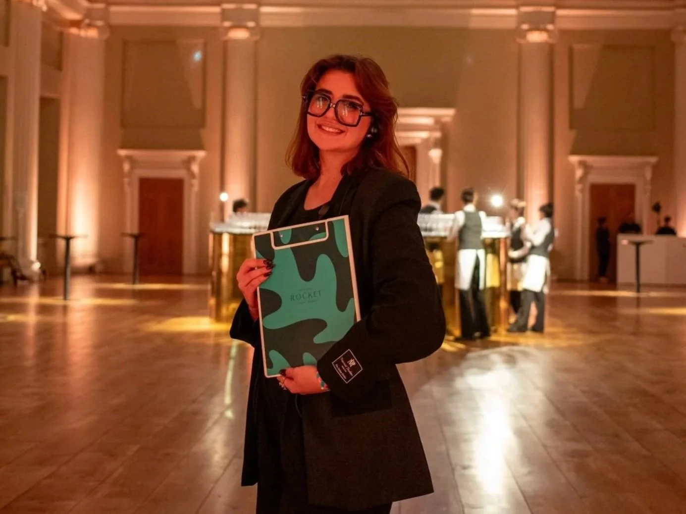A woman with long dark hair and bangs, wearing a black blazer, standing outdoors in a park with green trees and colorful flowers.