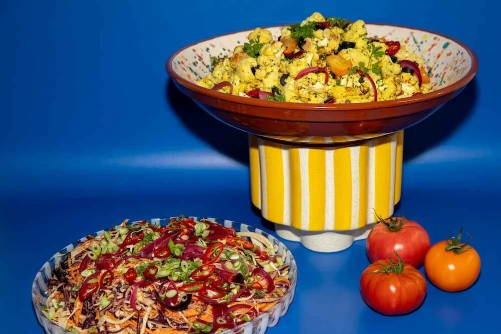 Two colorful bowls of salad, one on a striped yellow and white stand and the other on a blue surface, with three tomatoes beside them against a blue background.