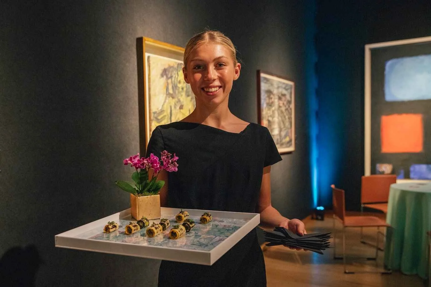 A smiling woman in a black dress holding a tray with a potted plant and wrapped candies, standing in an art gallery with paintings on the wall behind her.