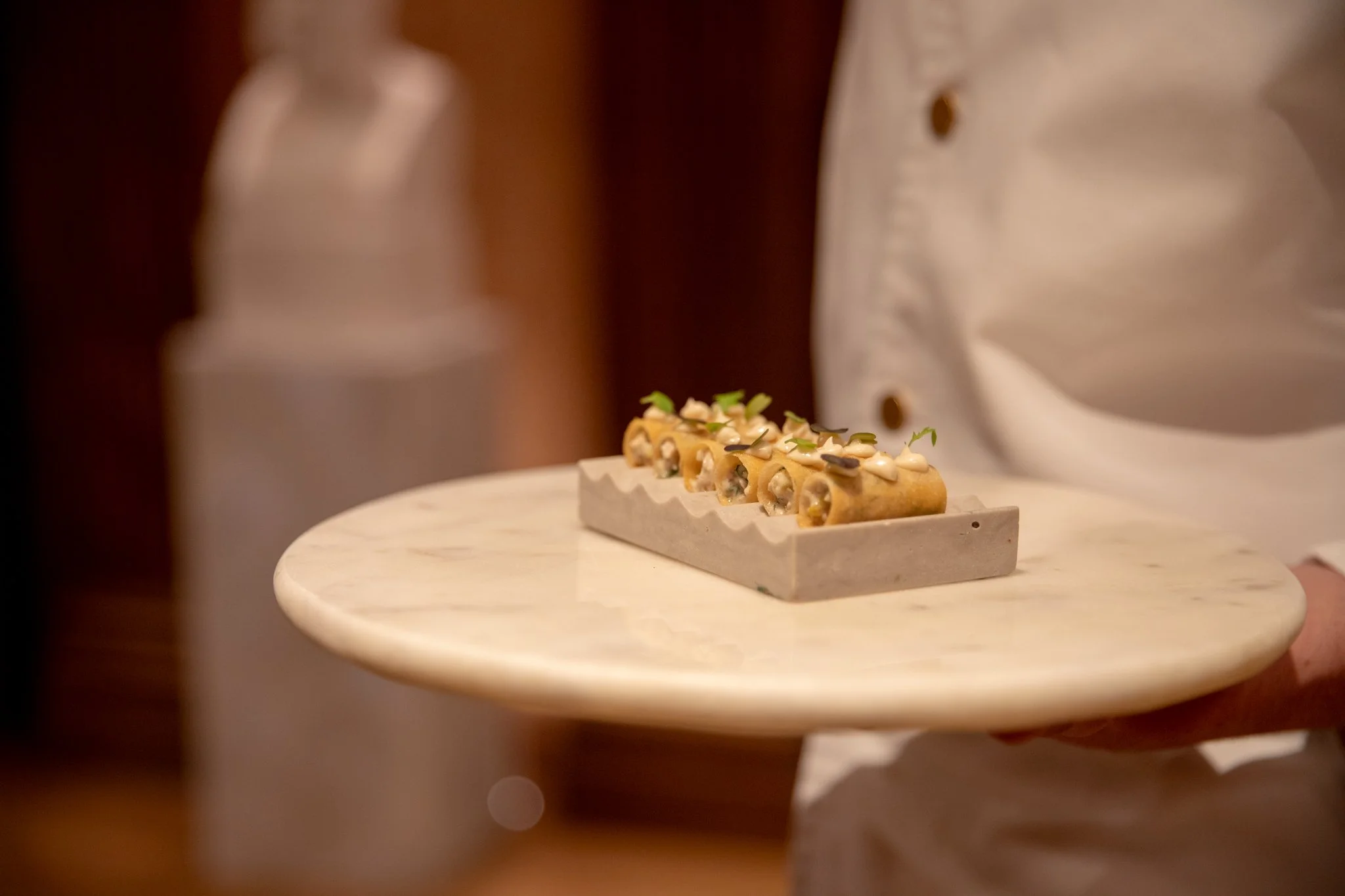 A person in a white chef's coat holding a round marble serving plate with four gourmet spring rolls garnished with microgreens, in a setting with warm lighting.